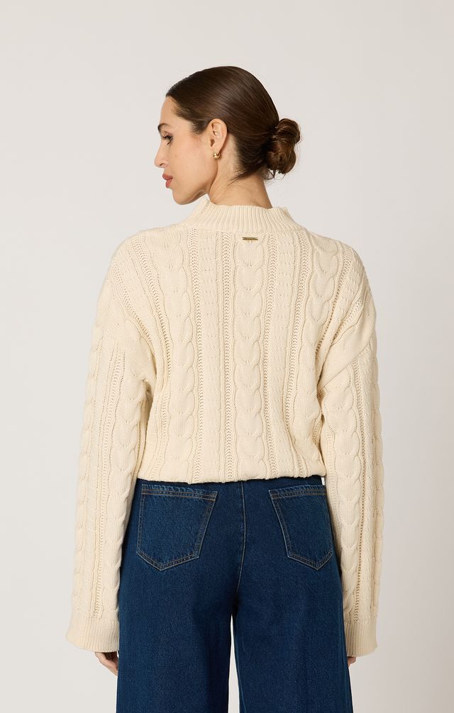 Woman wearing a cream cable knit cardigan and blue jeans on a plain background