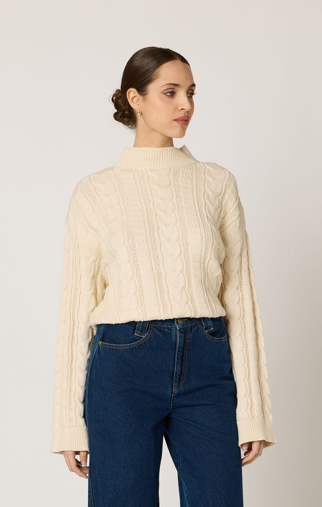 Woman wearing a cream cable knit sweater and blue jeans against a plain background