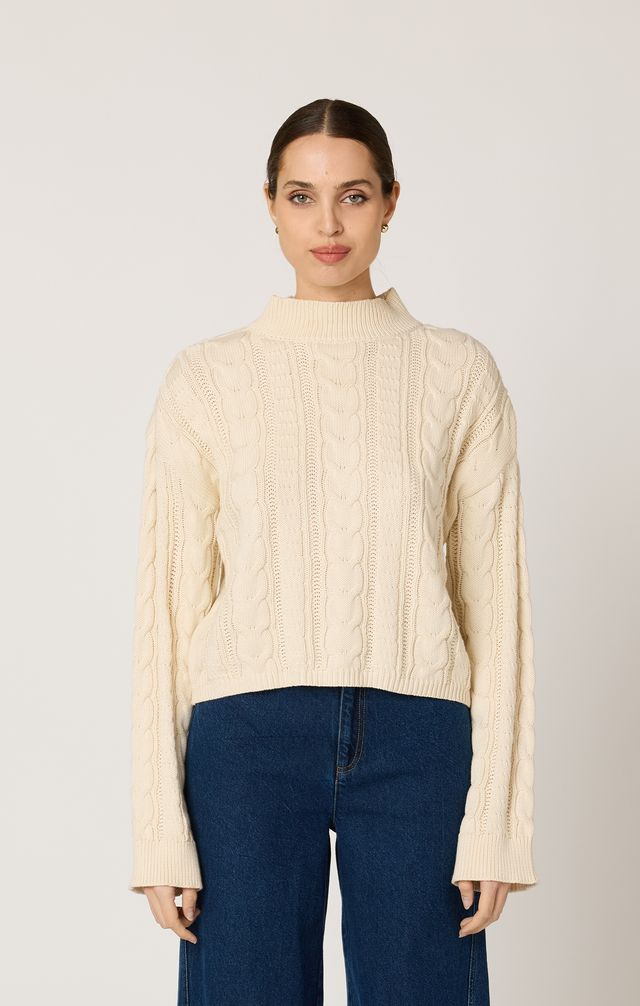 Woman wearing a beige cable knit sweater and blue jeans against a plain background
