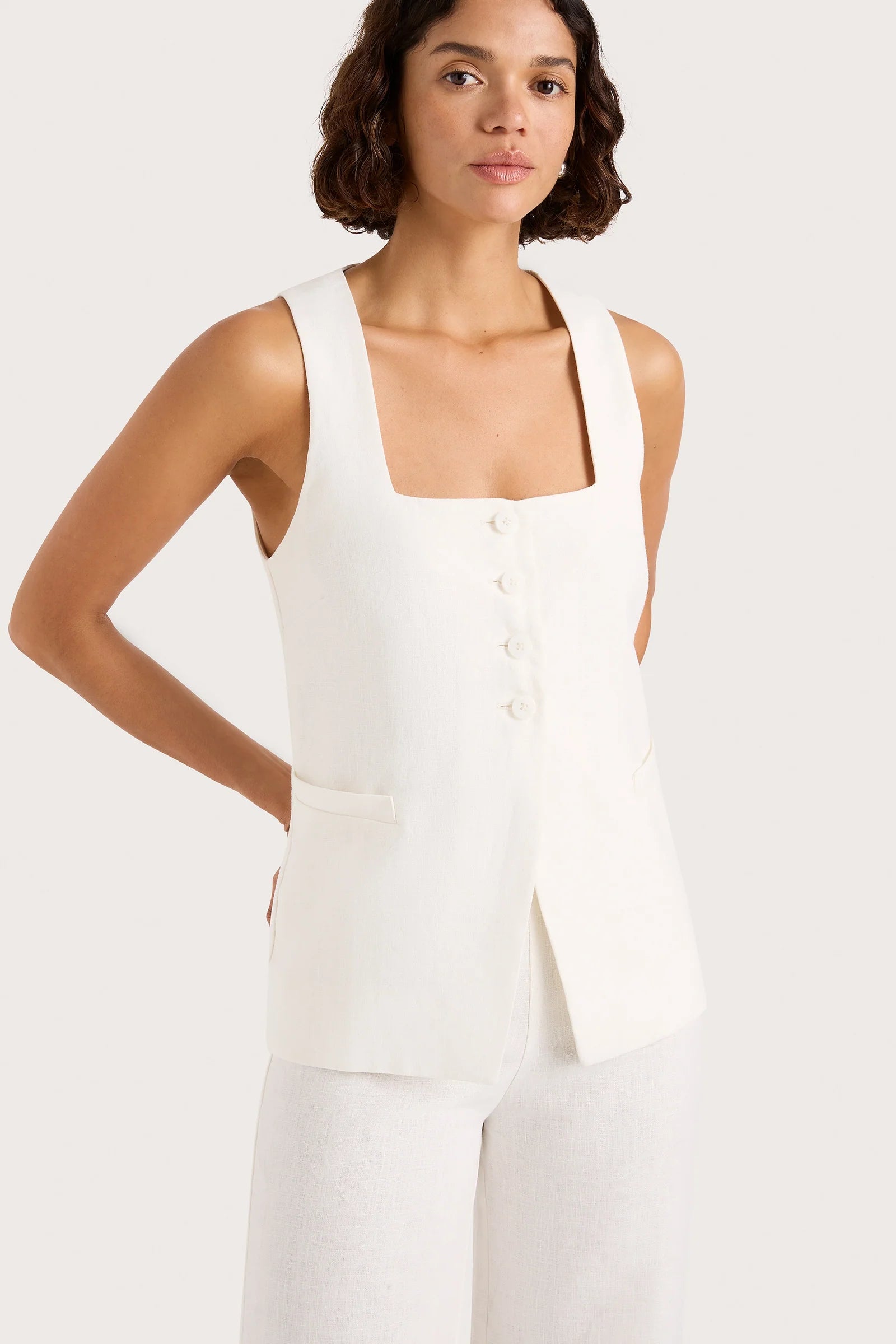Woman wearing a white sleeveless top with buttons on a plain background